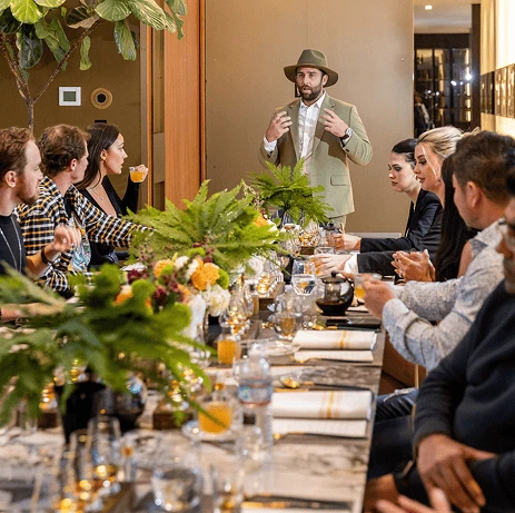 A group of people seated around a decorated dining table listening to a man in a green jacket and hat speaking at a formal gathering.