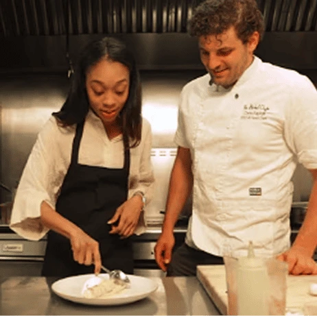 Chef instructing a woman in a kitchen as she prepares food on a plate.