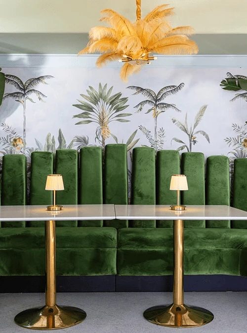 Stylish café interior with green velvet seating, gold tables, and a decorative feather chandelier against a tropical mural wall.
