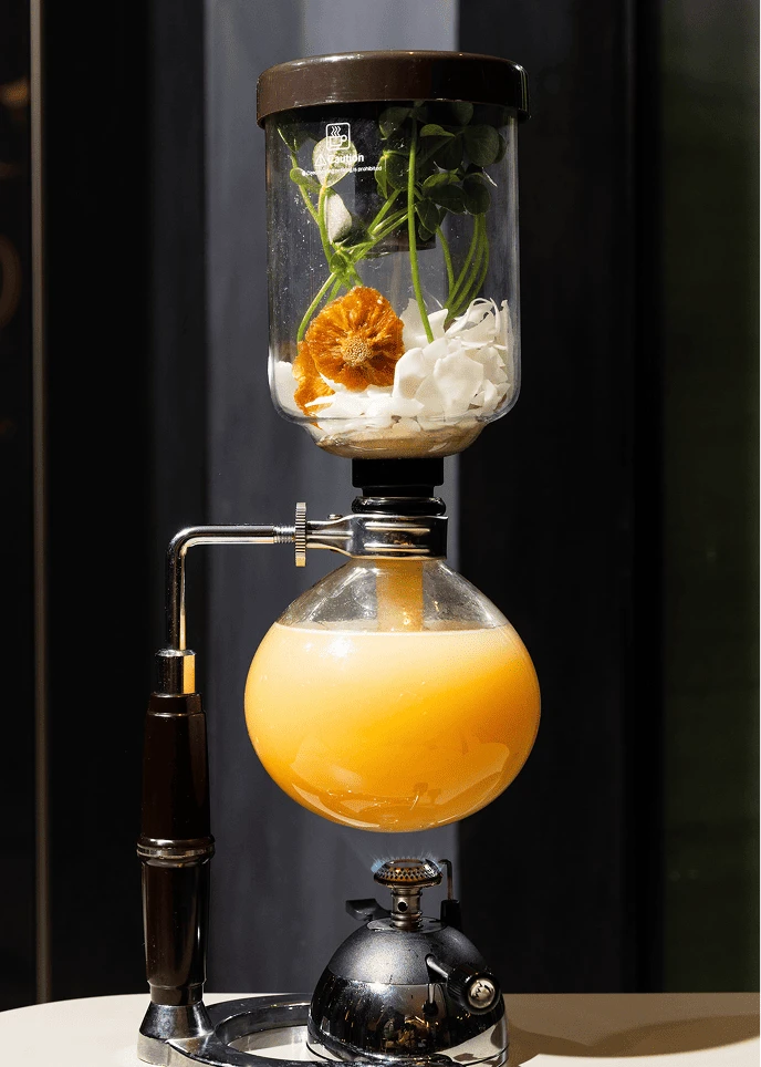 Glass siphon coffee maker filled with herbs, flowers, and orange liquid.