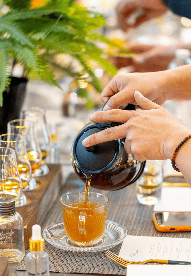 Close-up of a person pouring tea or a warm beverage into a glass cup during a tasting event, with bottles, glasses, and notes on the table.