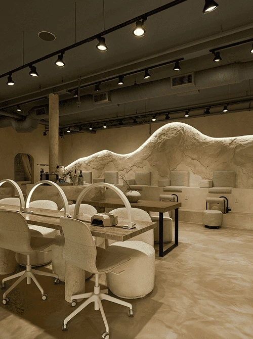 Modern beige-toned nail salon interior with manicure stations, chairs, and artistic wavy wall design under track lighting.