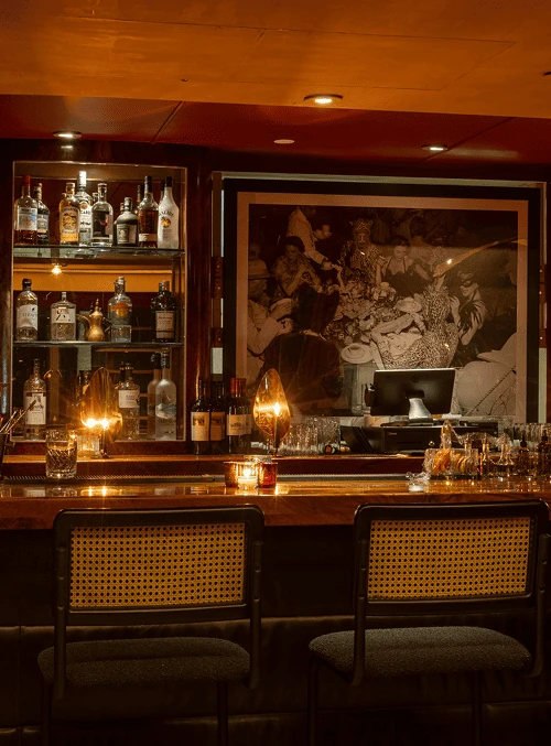 Cozy bar interior with shelves of liquor bottles, warm lighting, and two chairs at the counter.