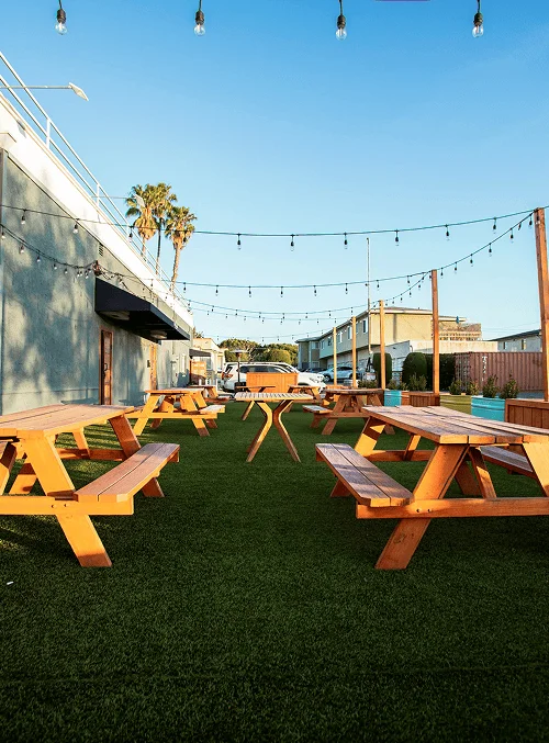 Outdoor seating area with wooden picnic tables on artificial grass, decorated with hanging string lights.