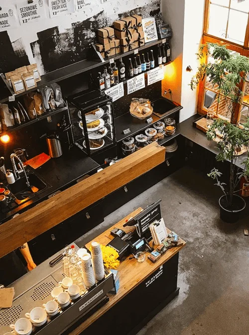Cozy modern café interior with a coffee counter, shelves stocked with products, and warm natural light from a window.