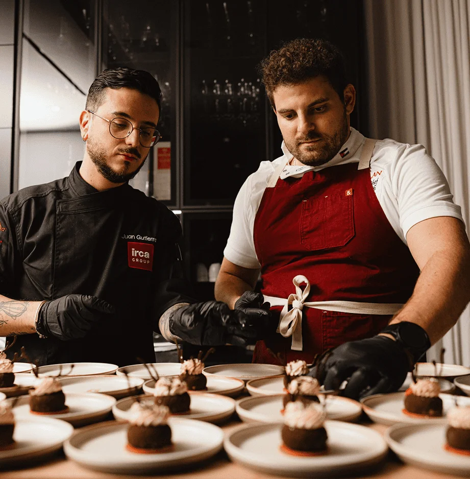 Two chefs plating desserts on multiple plates in a kitchen.