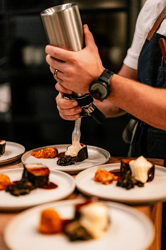 Chef plating dessert by piping cream onto a dish with several plates arranged on a counter.