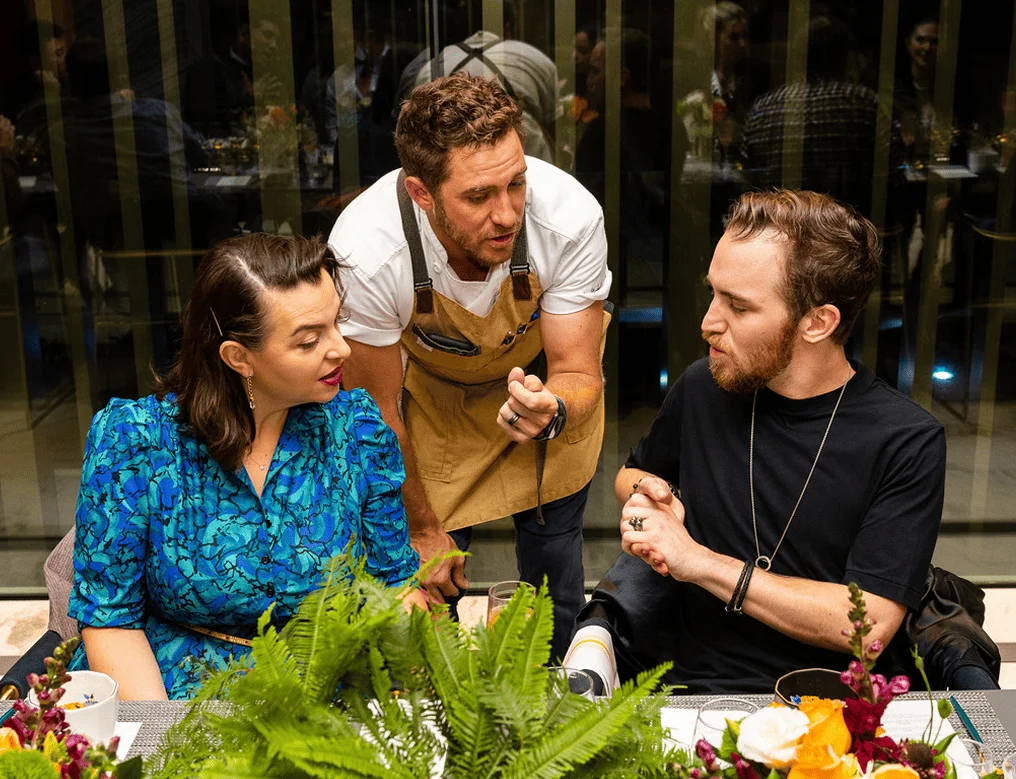 Chef interacting with two guests seated at a decorated dining table during a catered event.